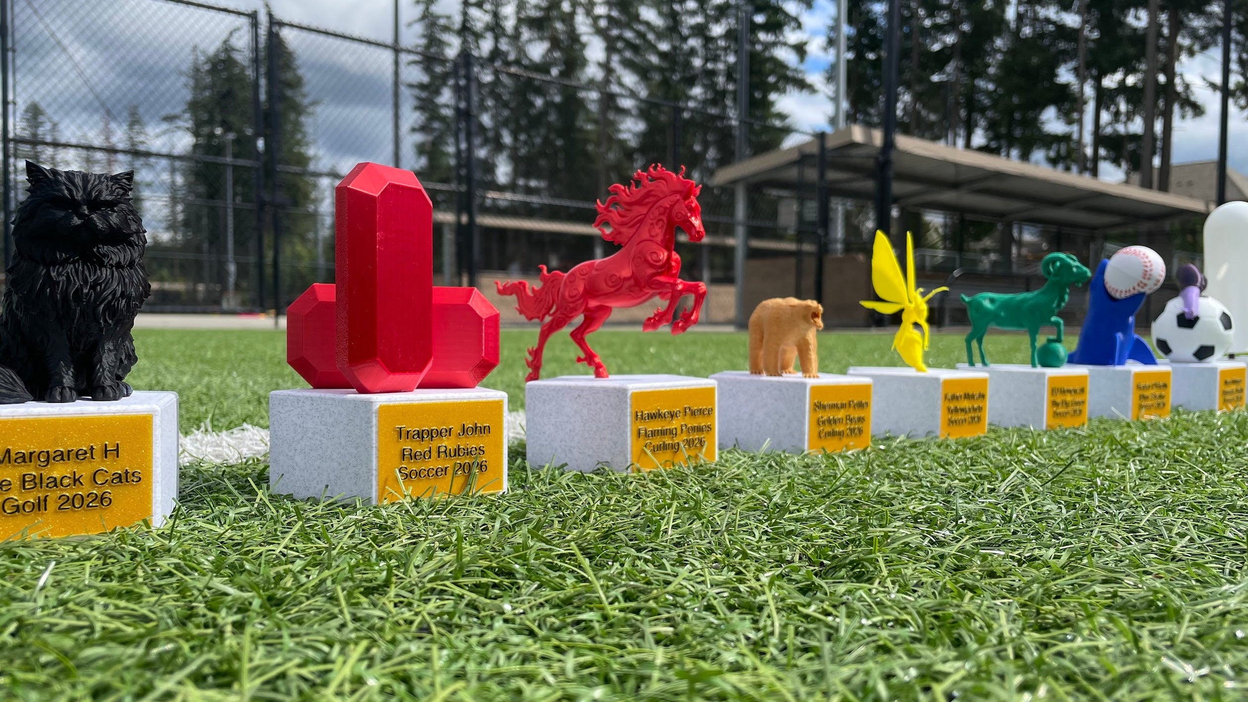 A row of colorful trophies displayed on white pedestals with yellow nameplates on green turf, with a chain-link fence and trees in the background.